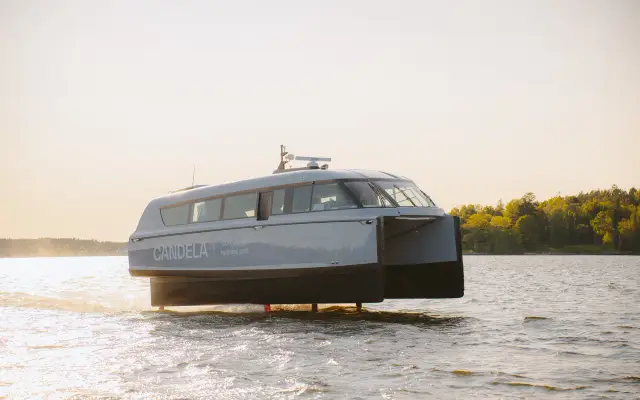 A white electric foiling ferry on a lake, hull lifted on foils, with mountains behind it.
